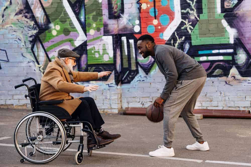 Old caucasian man in a wheelchair playing hobby basketball with an able bodied young black male