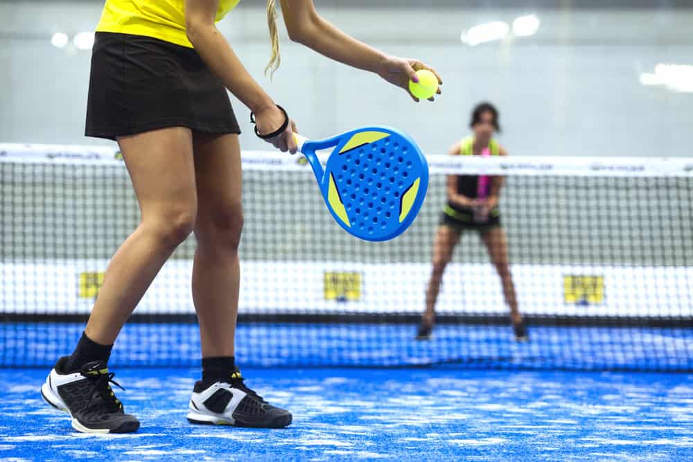 Women playing pickleball with paddle and ball on indoor court.