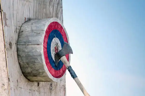 An axe embedded in the bullseye of a red, white, and blue wooden axe-throwing target mounted on a wood slab, shot outdoors against a clear sky.