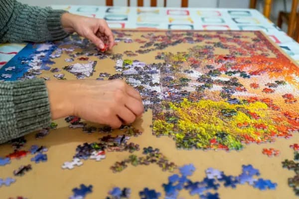 Close-up of a woman's hand in a grey knit sweater placing a puzzle piece into a large, colorful jigsaw puzzle spread across a wooden table.