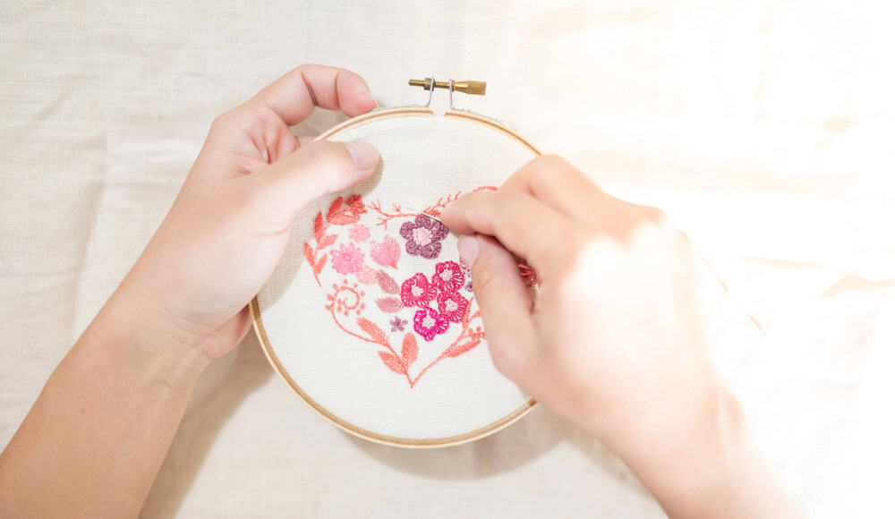 Two hands holding a wooden embroidery hoop, stitching a pink and magenta floral pattern onto cream fabric