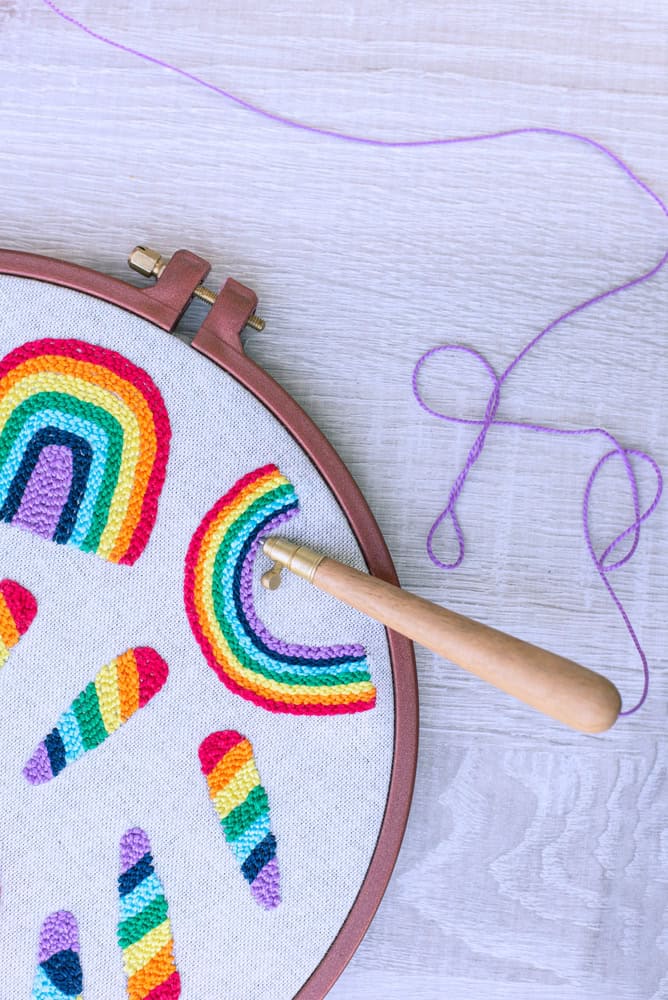 Close-up of a hand punch needling a rainbow design with colorful yarn into fabric stretched in an embroidery hoop